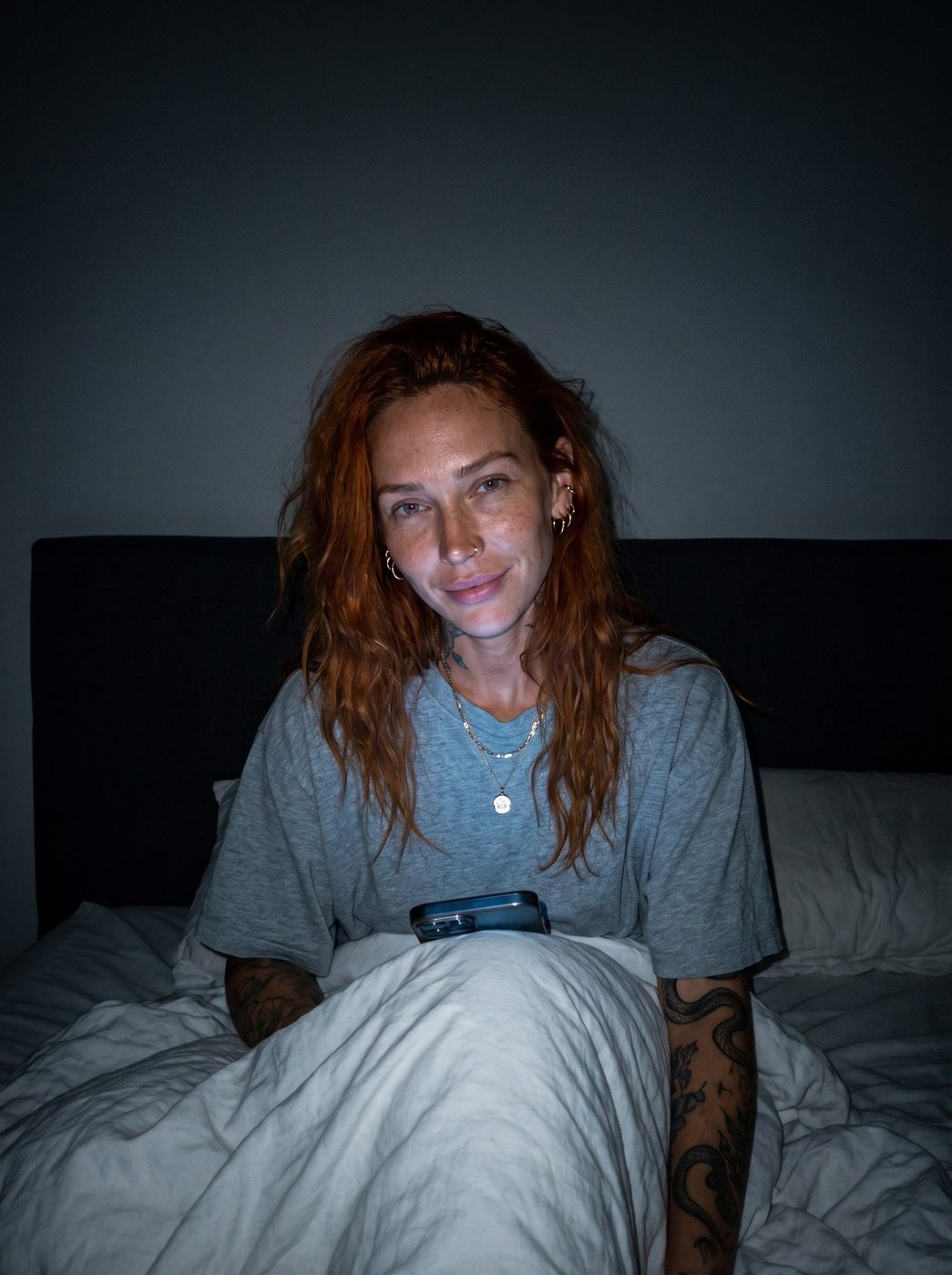 Ruby sitting up in bed at night in a grey T-shirt, the duvet over her knees and her phone lying on it, looking at the camera
